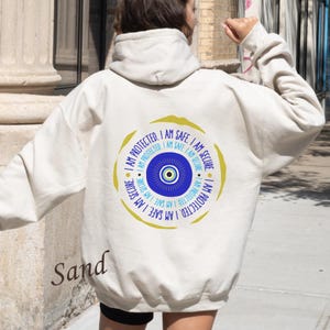 May include: A white hoodie with a large, gold circle design featuring a blue evil eye with white and light blue accents. The circle has the text "I am protected. I am safe. I am secure." repeated around the edge.