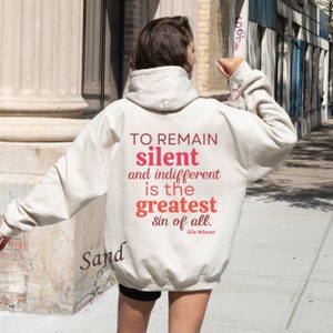 May include: A white hoodie with a quote printed on the back: "TO REMAIN silent and indifferent is the greatest sin of all. Elie Wiesel". The word "silent" is in red and the rest of the text is in pink.