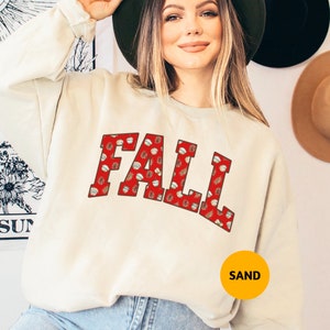 May include: A sand-coloured sweatshirt featuring the word "FALL" in a red, collegiate-style font. The letters are filled with a pattern of acorns and baseballs. This casual sweatshirt has a fall theme.