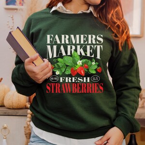 May include: Green sweatshirt with the text "Farmers Market" and "Fresh Strawberries" in red. The text "You Pick" and "$1.25" are also included in smaller font. A graphic of strawberries and leaves is featured.