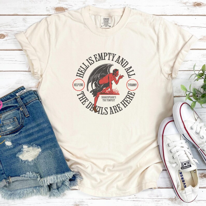 Puede incluir: Camiseta color crema con un gr&aacute;fico de diablo rojo y el texto "HELL IS EMPTY AND ALL THE DEVILS ARE HERE." El dise&ntilde;o incluye las frases "HELP END" y "TYRANNY." La camiseta se combina con pantalones cortos de mezclilla y zapatillas blancas.