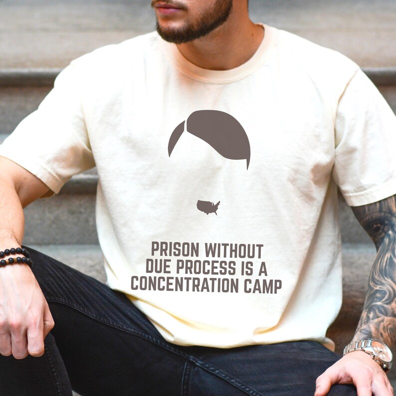 May include: Cream-colored t-shirt featuring a minimalist graphic of a head and a map of the United States. The text on the shirt reads "PRISON WITHOUT DUE PROCESS IS A CONCENTRATION CAMP."