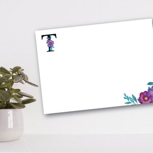 Puede incluir: Una tarjeta blanca con una letra negra "T" decorada con flores moradas y hojas azules. La letra está en la esquina superior izquierda de la tarjeta. La esquina inferior derecha de la tarjeta tiene un grupo de flores moradas y hojas azules.