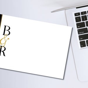Può includere: Una carta bianca con le lettere "B & R" in nero e oro, accanto a un laptop. La carta ha un bordo nero con un accento dorato. Il laptop è argentato e ha una tastiera nera.