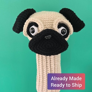 May include: A beige and black crocheted pug dog headcover for a golf club. The headcover features large, googly eyes and a black snout and ears. The text "Already Made Ready to Ship" is on a purple and red label.