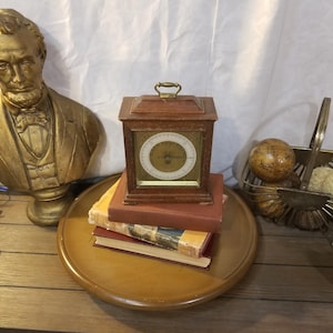 Puede incluir: Un barómetro de madera con asa de latón y esfera circular se encuentra sobre una pila de libros. También se ven un busto dorado y una cesta decorativa de metal con un globo.