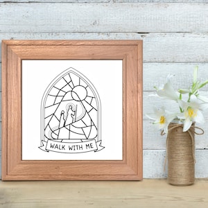 May include: A framed black and white print featuring a stained-glass style illustration of two figures and a sun. The banner at the bottom reads "WALK WITH ME". The frame is a light brown color. A vase with white flowers sits to the right.