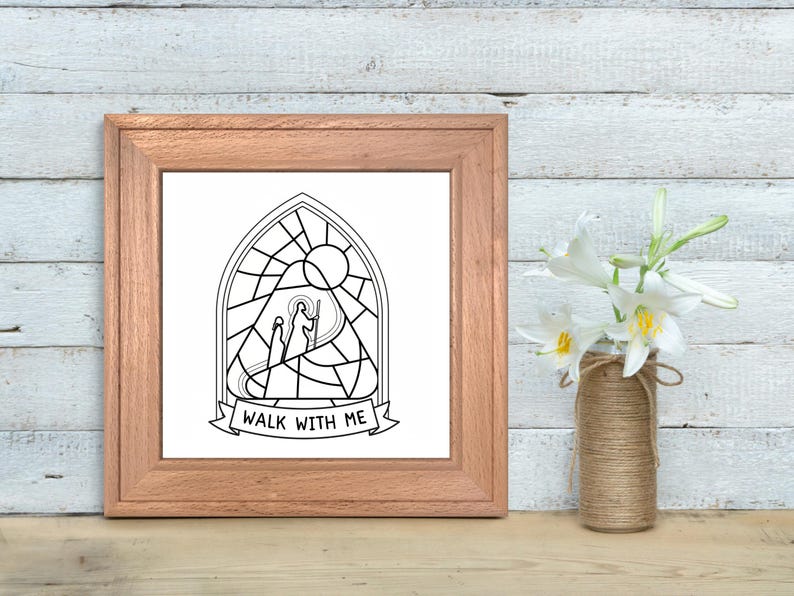 May include: A framed black and white print featuring a stained-glass style illustration of two figures and a sun. The banner at the bottom reads "WALK WITH ME". The frame is a light brown color. A vase with white flowers sits to the right.