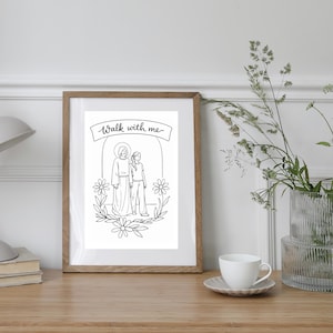May include: A framed black and white illustration with the text "Walk with me." The artwork depicts two figures, one with a halo, surrounded by floral elements. The frame is a light brown color, and the image is displayed on a wooden surface.