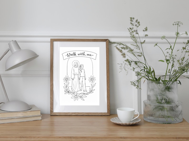 May include: A framed black and white illustration with the text "Walk with me." The artwork depicts two figures, one with a halo, surrounded by floral elements. The frame is a light brown color, and the image is displayed on a wooden surface.