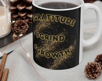 May include: A white ceramic mug with a black background and the words "#GRATITUDE", "#GRIND", and "#GROWTH" in gold lettering. The mug is filled with a dark liquid and has a white handle. The mug is on a light gray surface.