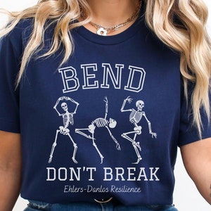 May include: A navy blue t-shirt with a white graphic of three skeletons dancing. The text "Bend Don't Break" is printed below the skeletons, with the words "Ehlers-Danlos Resilience" printed below that.