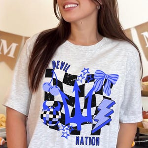 May include: A light gray t-shirt with a graphic design featuring a blue trident, cherries, stars, a bow, and a lightning bolt over a black and white checkered pattern. The words "DEVIL NATION" are also in the design.