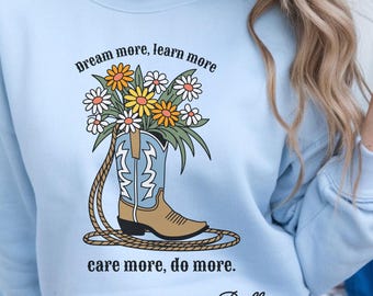 May include: Light blue sweatshirt with the text "Dream more, learn more, care more, do more." and "-Dolly." A graphic of a cowboy boot filled with flowers is in the center. The boot is tan and blue with a rope around it.
