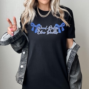 May include: Black t-shirt with the text "Wood-Ridge Blue Devils" in blue script and bow accents. The shirt is a crew neck and is worn with a denim jacket. The overall style is casual and sporty.