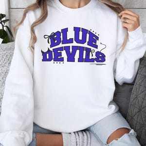 May include: A white long-sleeve crewneck sweatshirt featuring the text BLUE DEVILS in bold purple block letters. The design includes black decorative accents such as small bows, dots, and a devil tail extending from the letter S on the right side.