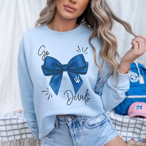 May include: A light blue long-sleeve sweatshirt featuring a large dark blue bow graphic in the center. The text "Go" is printed above the bow, and "Devils" is printed below it. A small white trident symbol is visible on the right side of the bow.