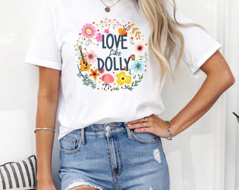 May include: White t-shirt with a floral wreath design and the text "Love Like Dolly." The wreath includes colorful flowers, leaves, and a small guitar. The model is wearing blue jeans.