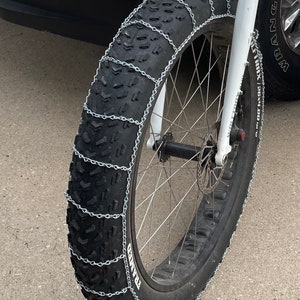 May include: A black fat bike tire with silver chains wrapped around it for traction. The tire is mounted on a white bike frame. The bike is parked on a concrete surface.