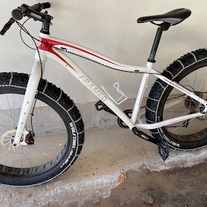 May include: White and red Framed fat tire bike with black tires. The bike has a black seat, handlebars, and pedals. The tires have chains wrapped around them. The bike has disc brakes and a water bottle cage.