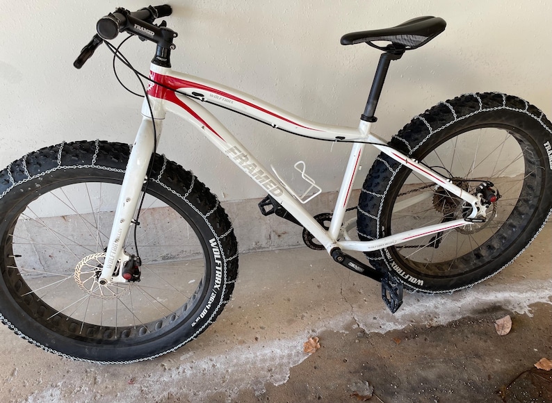May include: White and red Framed fat tire bike with black tires. The bike has a black seat, handlebars, and pedals. The tires have chains wrapped around them. The bike has disc brakes and a water bottle cage.