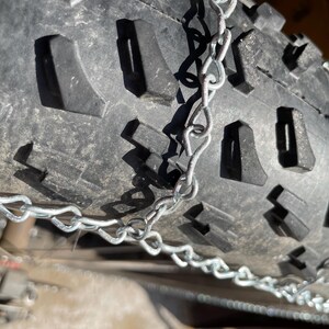 May include: Close-up of a black tire with a deep tread pattern and metal tire chains. The tire's surface is textured, and the chains are silver. The image is well-lit, highlighting the details of the tire and chains.