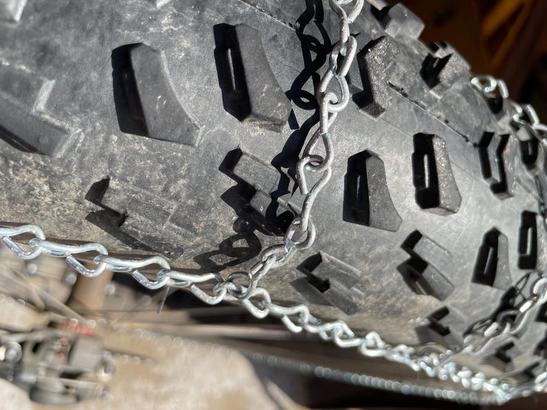 May include: Close-up of a black tire with a deep tread pattern and metal tire chains. The tire's surface is textured, and the chains are silver. The image is well-lit, highlighting the details of the tire and chains.