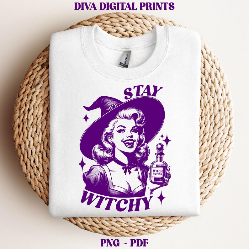 May include: White sweatshirt with a purple graphic of a smiling woman wearing a witch hat, holding a potion bottle. The text reads "STAY WITCHY" in purple. The sweatshirt is folded on a woven, circular surface.