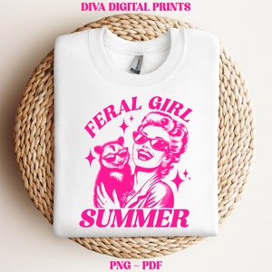 May include: A white t-shirt with a pink graphic design featuring a woman with sunglasses holding a possum. The text "Feral Girl Summer" is written in pink letters.