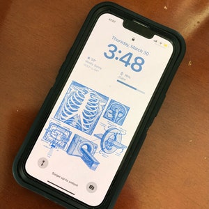 以下が含まれることがあります： 黒いケースに入ったスマートフォンは、青と白の医療イラストが描かれたロック画面を表示しています。画面には時刻、天気、バッテリー残量が表示されています。「スワイプしてロック解除」のテキストが表示されています。