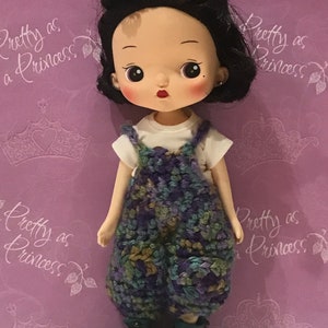 May include: A doll with black hair and a white shirt wearing crocheted overalls with green, purple, and brown accents. The doll is wearing green shoes with black accents and has a butterfly hair clip.