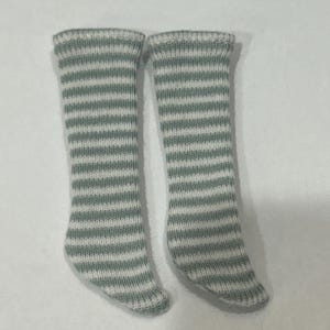 May include: A pair of knee-high socks with horizontal stripes in light green and white. The socks are knitted and appear to be made of a soft material. The socks are designed for a doll or small child.