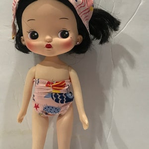 May include: A small doll with black hair and a pink and blue patterned swimsuit. The doll is wearing a pink and blue patterned bandana on her head.