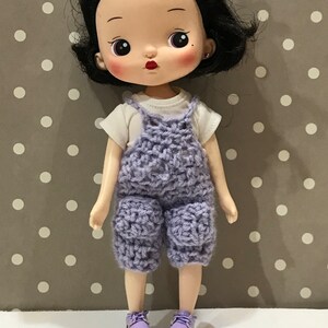 May include: A purple crocheted overall outfit with a white t-shirt and purple shoes on a doll with black hair and a light complexion.