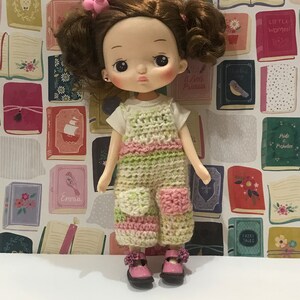 May include: A doll with brown hair and a pink crocheted dress. The doll is wearing pink shoes and has a pink bow in her hair. The doll is standing in front of a wall with a pattern of books.