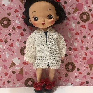 May include: A white crocheted cardigan with a button closure, worn by a doll with black hair and red shoes. The doll is standing against a pink background with a pattern of hearts, candy canes, and ice cream cones.