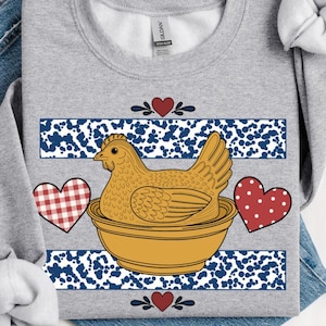 May include: A light gray sweatshirt featuring a hen in a bowl design. The hen is light brown, and the bowl is yellow. The design includes blue and white stripes, red hearts, and a gingham heart.