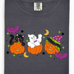 May include: Dark gray t-shirt featuring a Halloween design. The design includes a black cat, a ghost, and a jack-o'-lantern, each in a carved pumpkin. Purple bats, stars, and crescent moons complete the festive look.