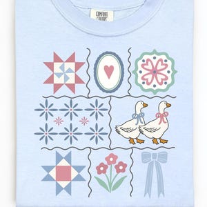 May include: Light blue sweatshirt with a patchwork design featuring floral and geometric patterns, two geese with bows, and a bow. The shirt has the Comfort Colors brand label.