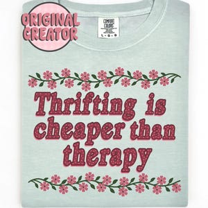 以下が含まれることがあります： 「Thrifting is cheaper than therapy」の文字が赤い水玉模様でプリントされた、薄い緑色のTシャツ。シャツには花のボーダーと、「Original Creator」の文字が入ったピンク色の円があしらわれています。