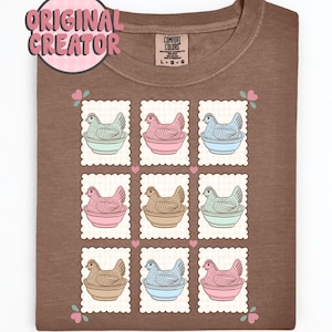 May include: Brown t-shirt featuring a grid of nine postage stamp-style illustrations. Each stamp depicts a hen in a bowl, with varying pastel colors. The shirt has a tag that says "Comfort Colors" and a pink "Original Creator" logo.