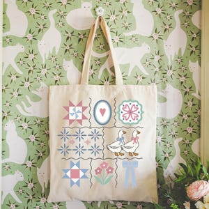 May include: A cream-colored tote bag with a patchwork design featuring geometric shapes, flowers, and geese. The bag hangs against a green floral wallpaper with white cat silhouettes. The tote bag has two handles.