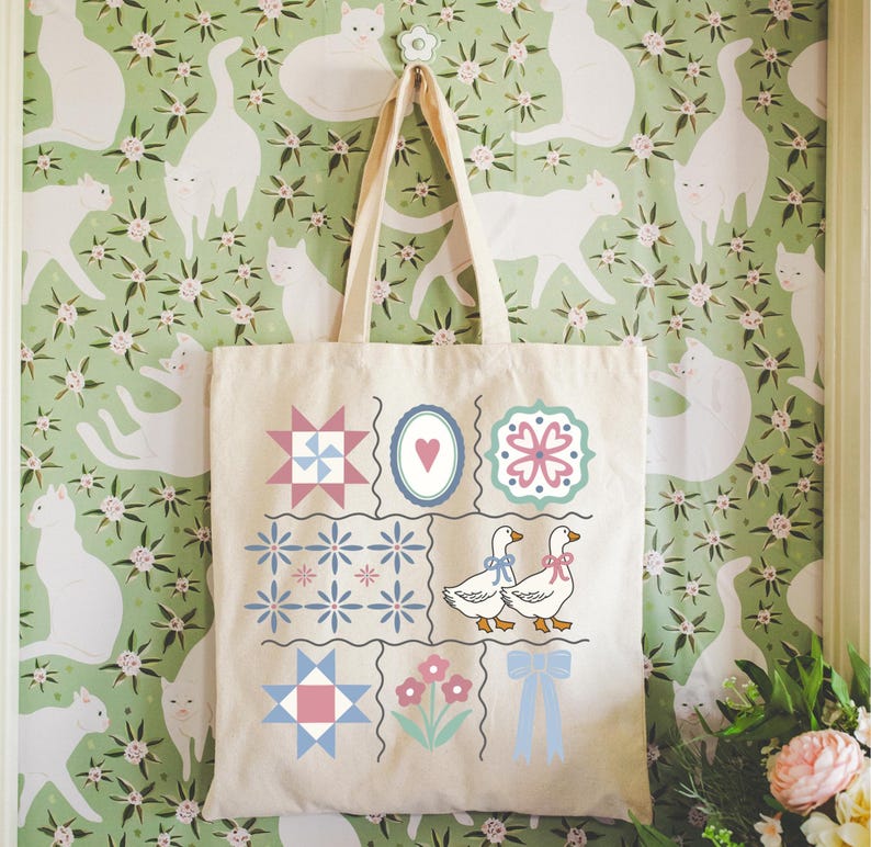 May include: A cream-colored tote bag with a patchwork design featuring geometric shapes, flowers, and geese. The bag hangs against a green floral wallpaper with white cat silhouettes. The tote bag has two handles.