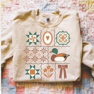 May include: Beige sweatshirt with a patchwork design featuring a duck, heart, flowers, and geometric shapes in green, brown, and orange. The sweatshirt has a crew neck and long sleeves, perfect for casual wear.