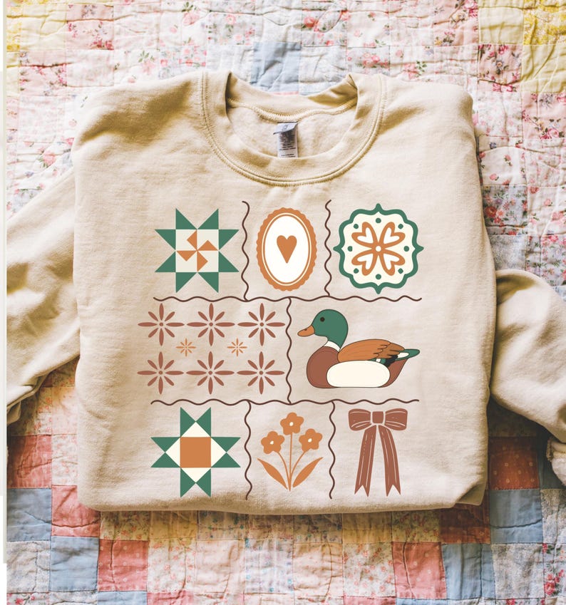 May include: Beige sweatshirt with a patchwork design featuring a duck, heart, flowers, and geometric shapes in green, brown, and orange. The sweatshirt has a crew neck and long sleeves, perfect for casual wear.