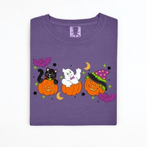 May include: Purple Comfort Colors t-shirt featuring a Halloween design. The design includes a black cat, a ghost, and a jack-o'-lantern, each in a carved pumpkin. Bats, stars, and moons complete the festive look.