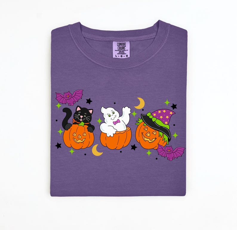 May include: Purple Comfort Colors t-shirt featuring a Halloween design. The design includes a black cat, a ghost, and a jack-o'-lantern, each in a carved pumpkin. Bats, stars, and moons complete the festive look.