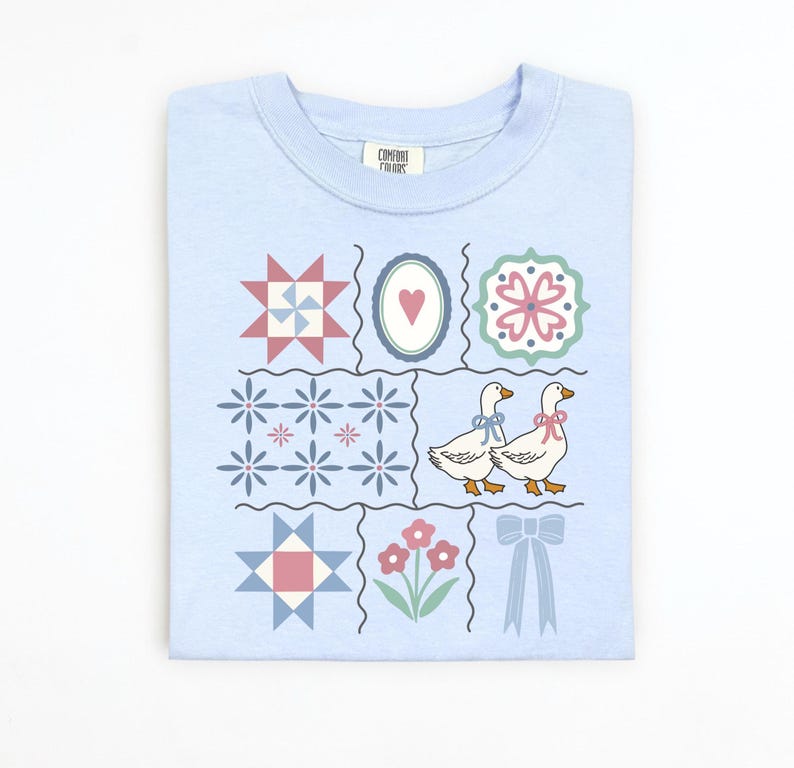 May include: Light blue sweatshirt with a patchwork design featuring floral and geometric patterns, two geese with bows, and a bow. The shirt has the Comfort Colors brand label.