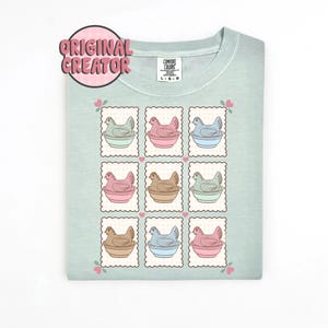 May include: A light green t-shirt featuring a grid of nine postage stamp-style illustrations. Each stamp depicts a hen in a bowl, with varying colors of pink, blue, brown, and green. The shirt has the text "ORIGINAL CREATOR" in a pink circle.