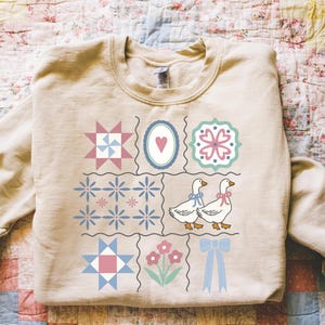 May include: A beige sweatshirt featuring a patchwork design with various quilt block patterns, including stars, flowers, and hearts. Two geese with blue and pink bows are also featured. The sweatshirt has a crew neck.
